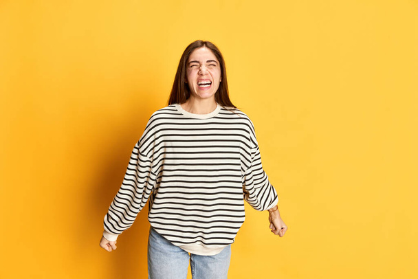 Portrait of young irritated girl in striped sweater emotionally showing anger, shouting against pink studio background. Concept of youth, emotions, facial expression, lifestyle - Φωτογραφία, εικόνα