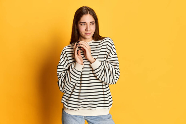 Portrait of young beautiful girl in striped sweater with plotting face posing against yellow studio background. Making plan. Concept of youth, emotions, facial expression, lifestyle - Foto, Bild