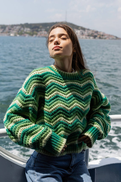Young woman in knitted sweater standing on yacht with sea and Princess islands at background in Turkey  - Fotó, kép