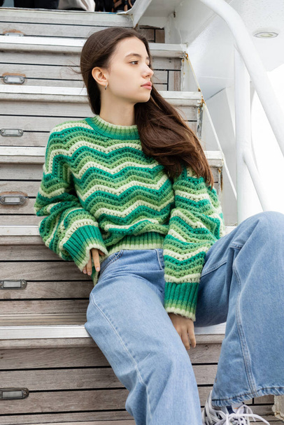Young brunette woman in sweater and jeans sitting on stairs on yacht  - Foto, afbeelding