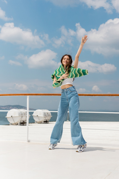 Cheerful young woman in sweater and jeans waving hand while standing on ferry boat in Turkey  - Photo, Image