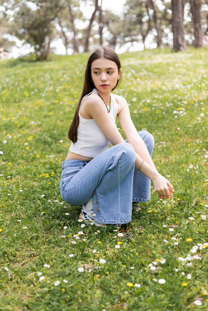 Young woman in top and jeans sitting on lawn with daisies at daytime  - Foto, Imagen