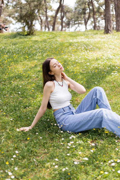 Jovem mulher em cima e jeans tocando pescoço enquanto sentado no gramado com flores no parque  - Foto, Imagem