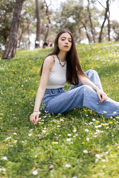Młoda kobieta w ubraniu casual patrząc w aparat podczas siedzenia na łące z kwiatami w parku - Zdjęcie, obraz
