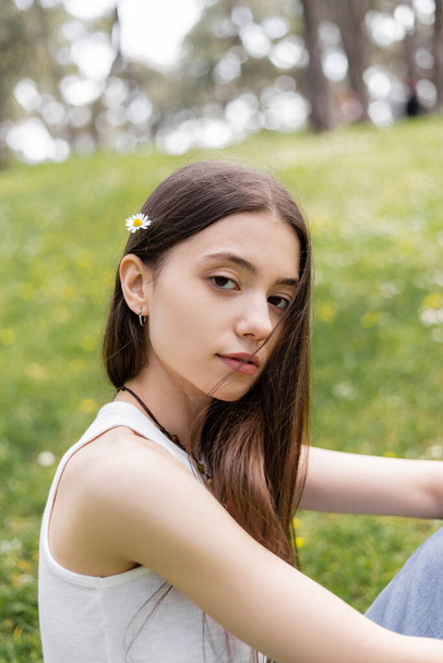 Portrait of brunette woman in top with daisy in hair looking at camera in park  - Photo, Image