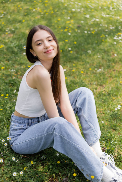 Overjoyed woman in top and jeans looking at camera while sitting on lawn with flowers in park  - Valokuva, kuva