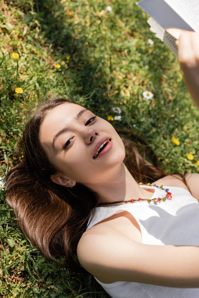 Vista de ángulo alto de la joven sonriente mirando a la cámara mientras sostiene el libro y yace en el prado con flores en el parque  - Foto, imagen
