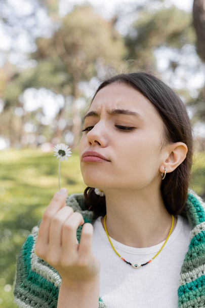 Konzentrierte junge Frau blickt auf Gänseblümchen-Blume im verschwommenen Sommerpark  - Foto, Bild