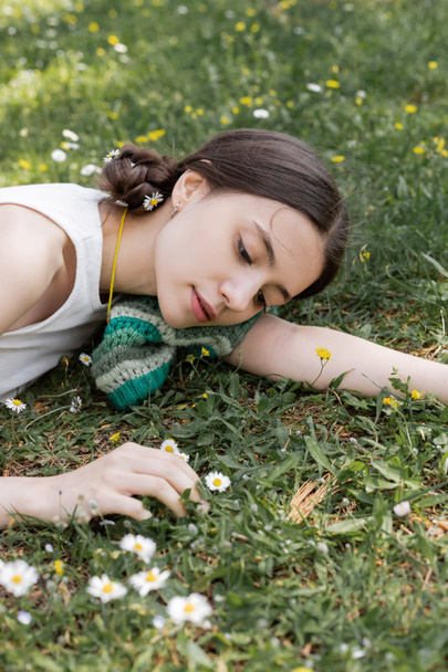 Retrato de una joven morena mirando margaritas borrosas mientras yacía en el prado en el parque  - Foto, Imagen