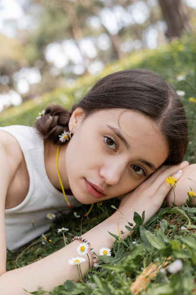 Retrato de una mujer joven y bonita mirando a la cámara mientras yacía en el prado con flores en el parque  - Foto, Imagen