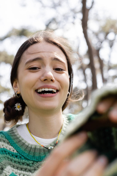 Mujer alegre con margarita en el pelo mirando a la cámara y riendo en el parque  - Foto, Imagen