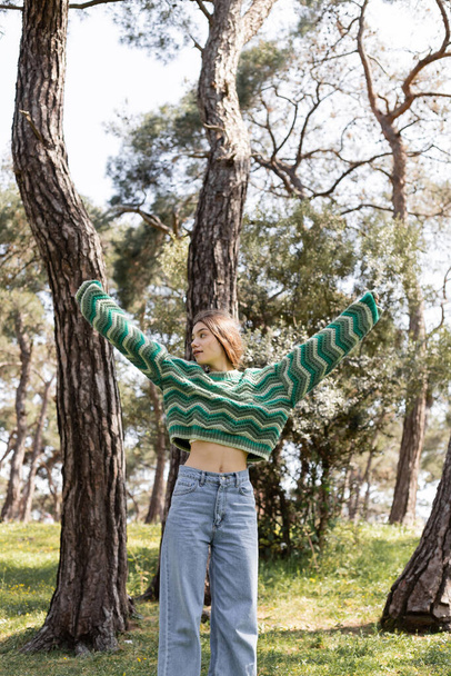 Jovem mulher em camisola de malha e jeans olhando para longe enquanto estava em pé no parque de verão  - Foto, Imagem
