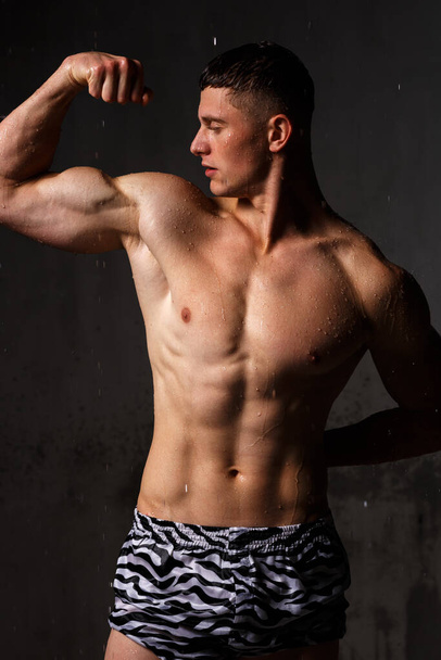 Bodybuilder επιδεικνύει τους μυς του και την εικόνα στο στούντιο στη βροχή. - Φωτογραφία, εικόνα