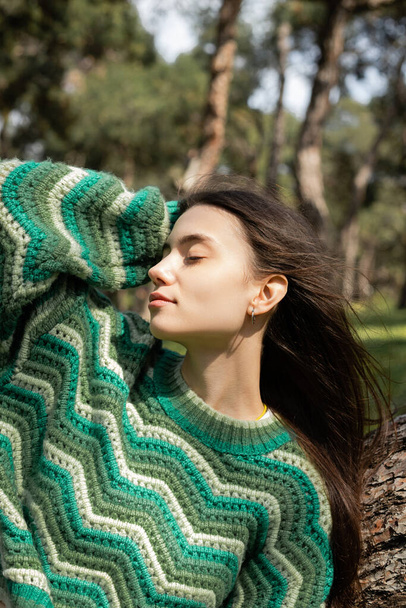 Young woman in knitted sweater touching hair near tree in park  - Fotoğraf, Görsel