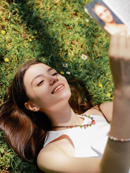 High angle view of smiling brunette woman reading book while lying on lawn with flowers  - Foto, Imagem