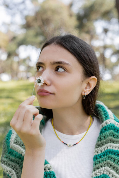 Young brunette woman smelling daisy and looking away in park  - Foto, Bild