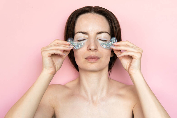 Pleased brunet woman undergoes beauty procedures apply hydrogel patches under eyes to remove puffiness and wrinkles isolated over pink background. Skin care beauty treatment at home - Fotografie, Obrázek