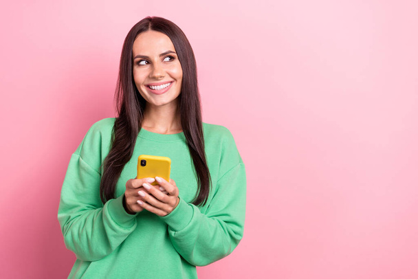 Photo of cheerful satisfied woman dressed green sweatshirt look at empty space holding smartphone isolated on pink color background. - Фото, изображение