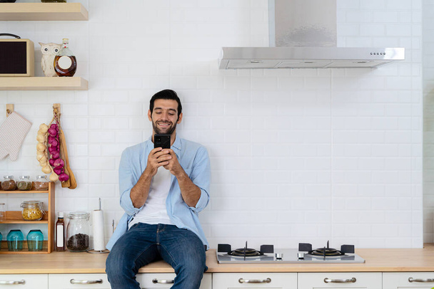 Handsome young man happy while sitting at home kitchen in good morning on leisurely vacation. Cheerful young bearded man talking on - Zdjęcie, obraz