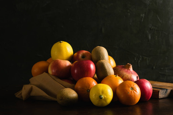 Fruit in a reusable bag. Earth day and zero waste concept. Orange, lemon, apple, kiwi, pomegranate and wooden cutting board on dark background. - 写真・画像
