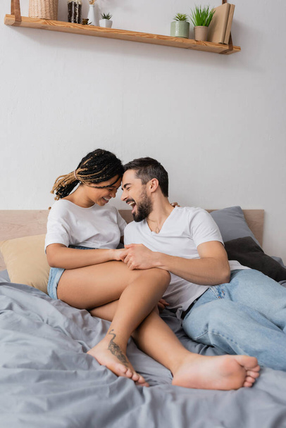 excited bearded man laughing near barefoot african american woman lying on bed at home - 写真・画像