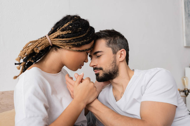 Sakallı adam ve genç Afrikalı Amerikalı kadın yatak odasında el ele tutuşuyorlar. - Fotoğraf, Görsel