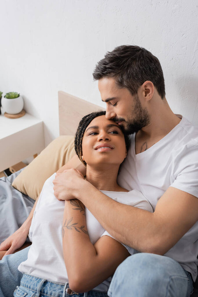 bebaarde man knuffelen jong en gelukkig Afrikaans amerikaanse vrouw glimlachen op bed thuis - Foto, afbeelding