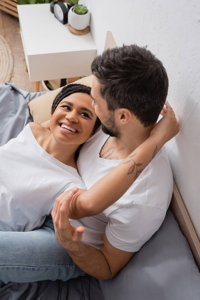 carefree african american woman in white t-shirt embracing boyfriend sitting on bed at home - Foto, Imagem