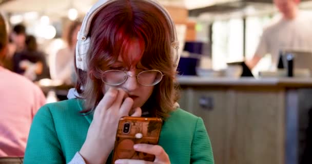  menina tenager com cabelo vermelho brilhante em óculos mordendo suas unhas e olhando para um gadget - Filmagem, Vídeo