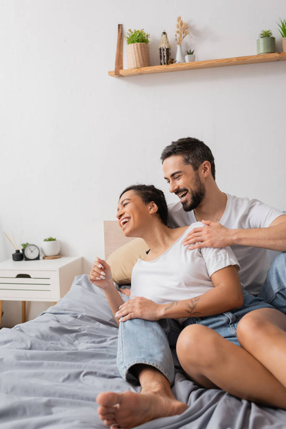 excited bearded man hugging cheerful african american woman while having fun on bed at home - 写真・画像