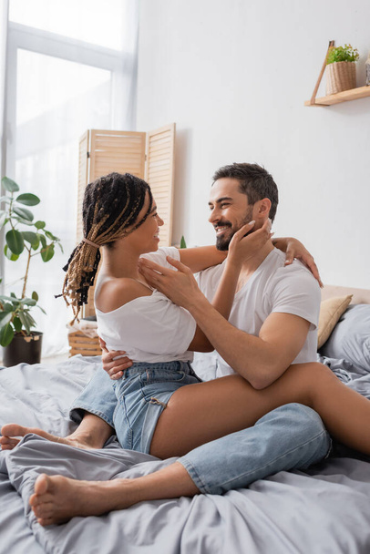 cheerful interracial couple in white t-shirts embracing and smiling at each other while sitting on bed at home - Photo, Image