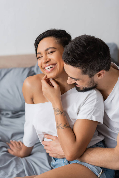 bearded man hugging carefree african american woman smiling with closed eyes in bedroom at home - Фото, изображение