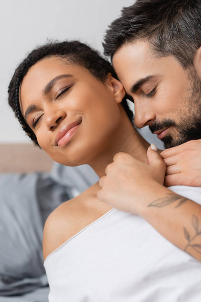 charming african american woman smiling with closed eyes while bearded man hugging her in bedroom at home - Valokuva, kuva