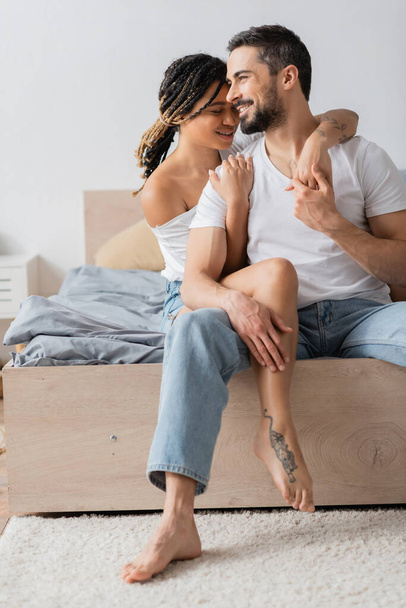 tattooed african american woman hugging happy bearded man sitting in jeans and white t-shirt on bed at home - Fotó, kép