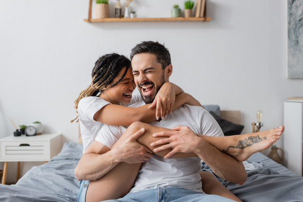 excited interracial couple in white t-shirts embracing while having fun in bedroom at home - Foto, imagen