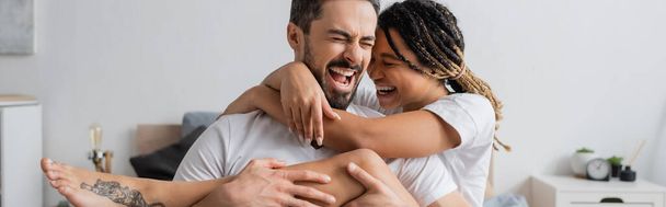 nadšený mezirasový pár v bílých tričkách objímající a smějící se se zavřenýma očima v ložnici doma, prapor - Fotografie, Obrázek