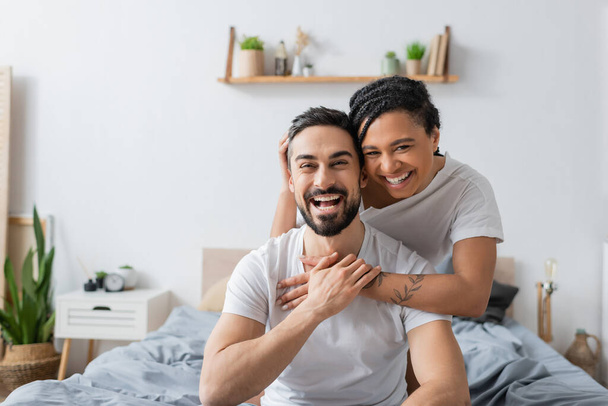 overjoyed multiethnic couple in white t-shirts laughing at camera in bedroom at home - Fotoğraf, Görsel