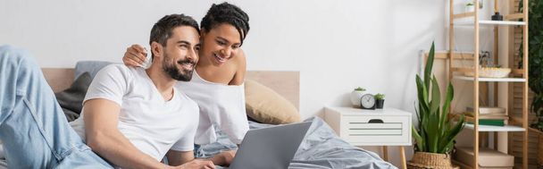 jong Afrikaans amerikaans vrouw en bebaarde man in wit t-shirts kijken film op laptop in slaapkamer, banner - Foto, afbeelding