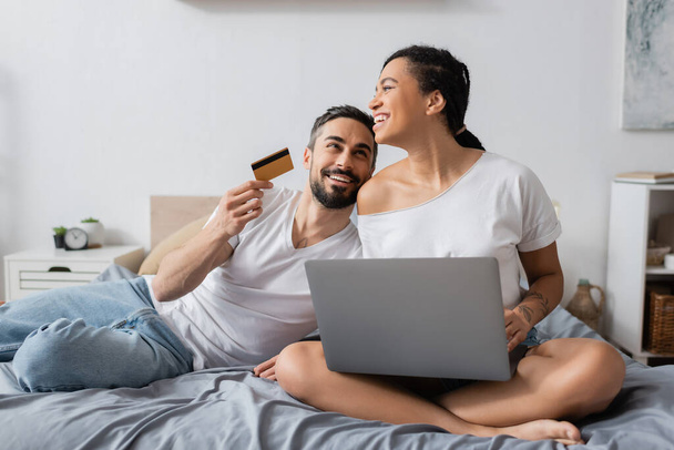 ευτυχισμένος γενειοφόρος άνδρας κρατώντας πιστωτική κάρτα κοντά γέλιο αφροαμερικανή φίλη κάθεται με φορητό υπολογιστή στο κρεβάτι στο σπίτι - Φωτογραφία, εικόνα