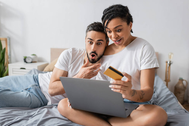 amazed african american woman with laptop and credit card near man with open mouth on bed at home - Foto, immagini