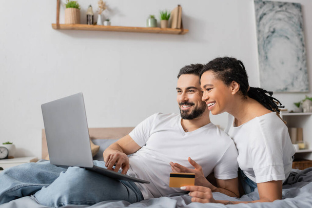 cheerful bearded man using laptop near african american woman with credit card on bed at home - Fotó, kép