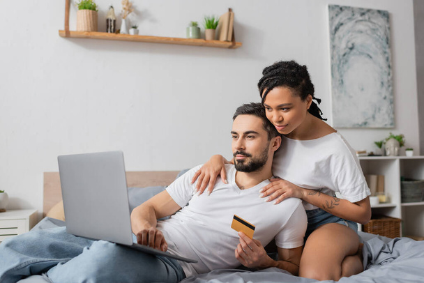 jeune femme afro-américaine étreignant les épaules de l'homme barbu avec ordinateur portable et carte de crédit sur le lit à la maison - Photo, image