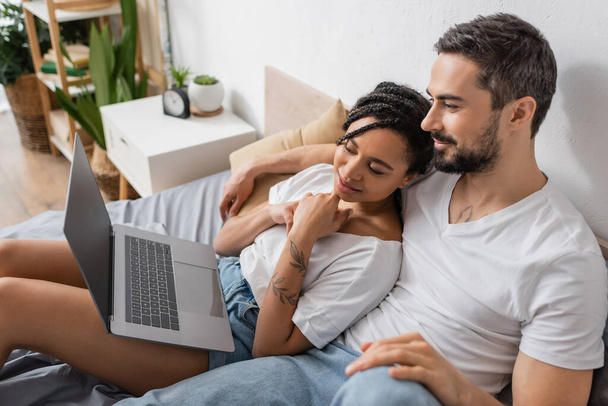 carefree interracial couple in white t-shirts watching movie on laptop in bedroom at home - Foto, immagini