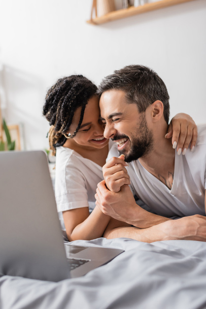 excited interracial couple laughing with closed eyes while watching comedy movie on laptop in bedroom - Fotografie, Obrázek