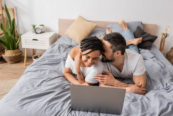 overhappy interraciale paar lachen met gesloten ogen terwijl het kijken van film op laptop in de slaapkamer thuis - Foto, afbeelding