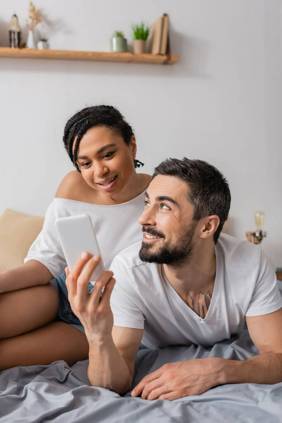 cheerful bearded man showing smartphone to smiling african american woman while resting on bed at home - Фото, изображение