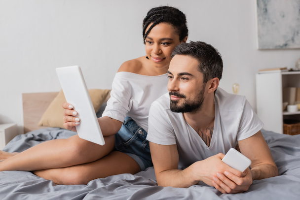 sorrindo mulher afro-americana mostrando tablet digital para namorado barbudo deitado com smartphone na cama - Foto, Imagem