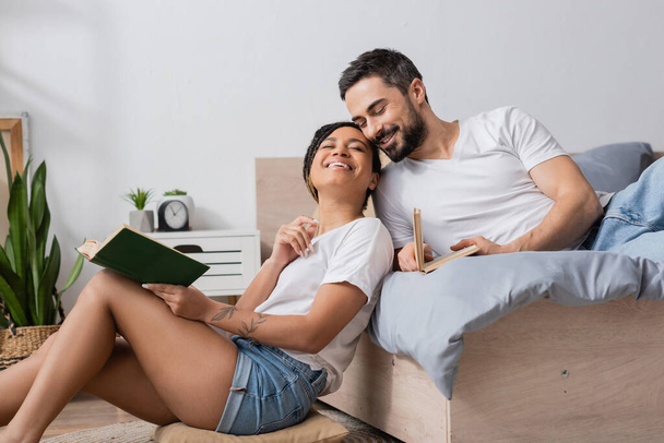 mulher afro-americana despreocupada com o livro sorrindo com os olhos fechados enquanto sentado perto do namorado sorridente deitado na cama em casa - Foto, Imagem