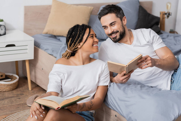 cheerful interracial couple in white t-shirts holding books and smiling at each other in bedroom at home - Φωτογραφία, εικόνα