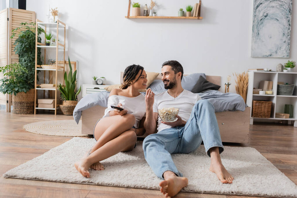 full length of barefoot and carefree interracial couple sitting with tv remote controller and popcorn on floor in modern bedroom - Foto, Bild